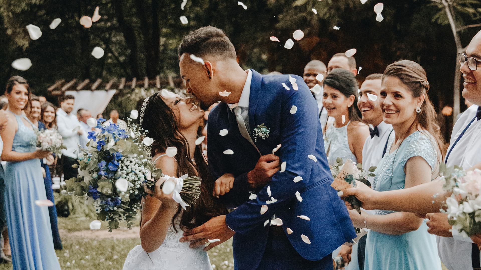 A picture to illustrate the top 10 Wedding Trends of 2024. This is a couple on their wedding day surrounded by their friends, family, and wedding party. Guests are throwing white and pink flowers into the air, and the couple is about to kiss as the flowers fall.
