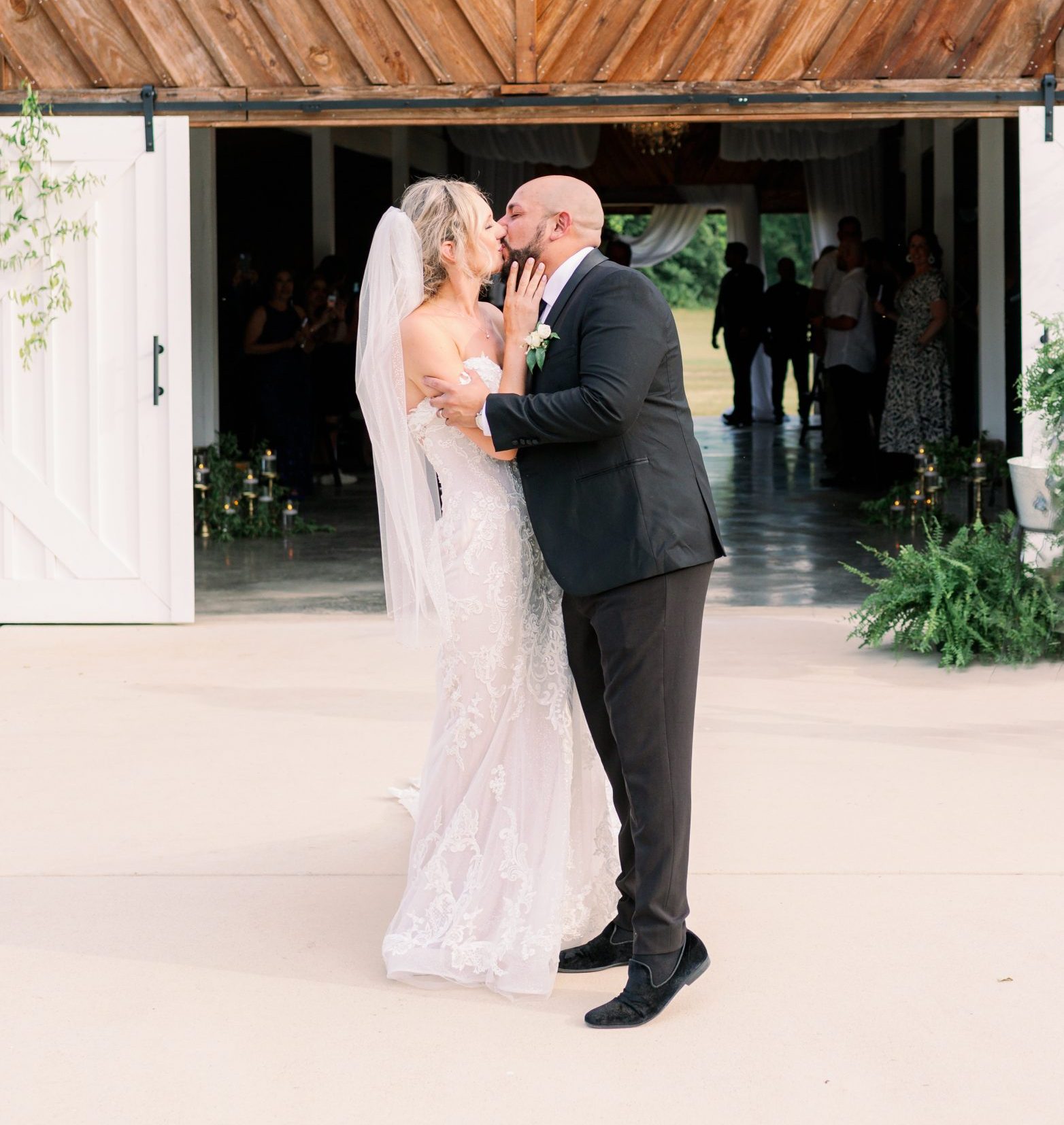 Bride and groom share a kiss in front of the barn's open doors, the bride's dress train spread out behind her on the concrete patio
