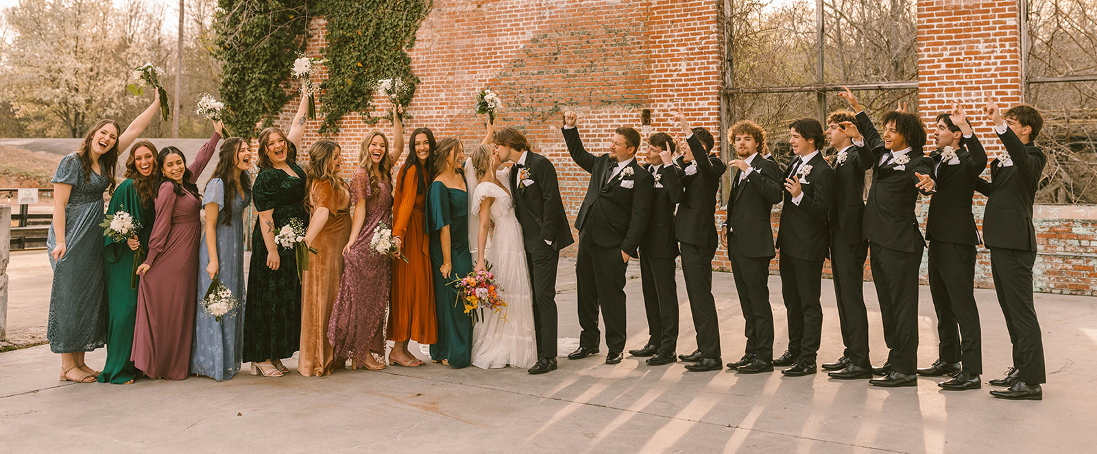 Colorful bridal party and groomsmen cheer as the bride and groom kiss in front of a historic brick building in Alabama.
