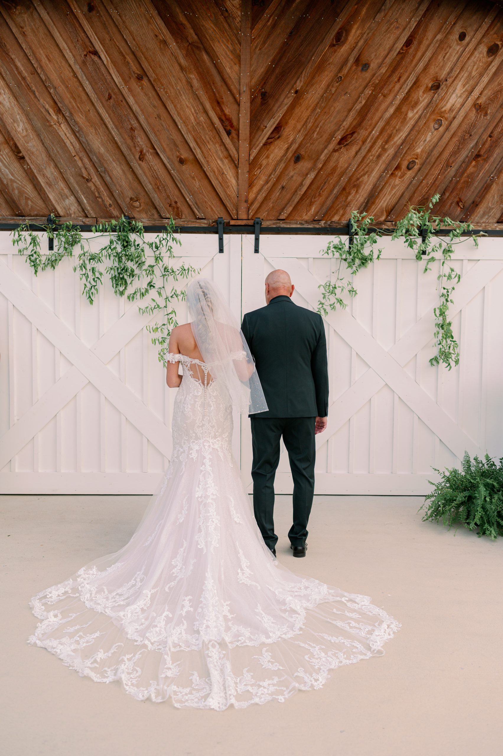Bride in lace wedding gown with her father standing before white barn doors at a rustic Huntsville wedding venue.