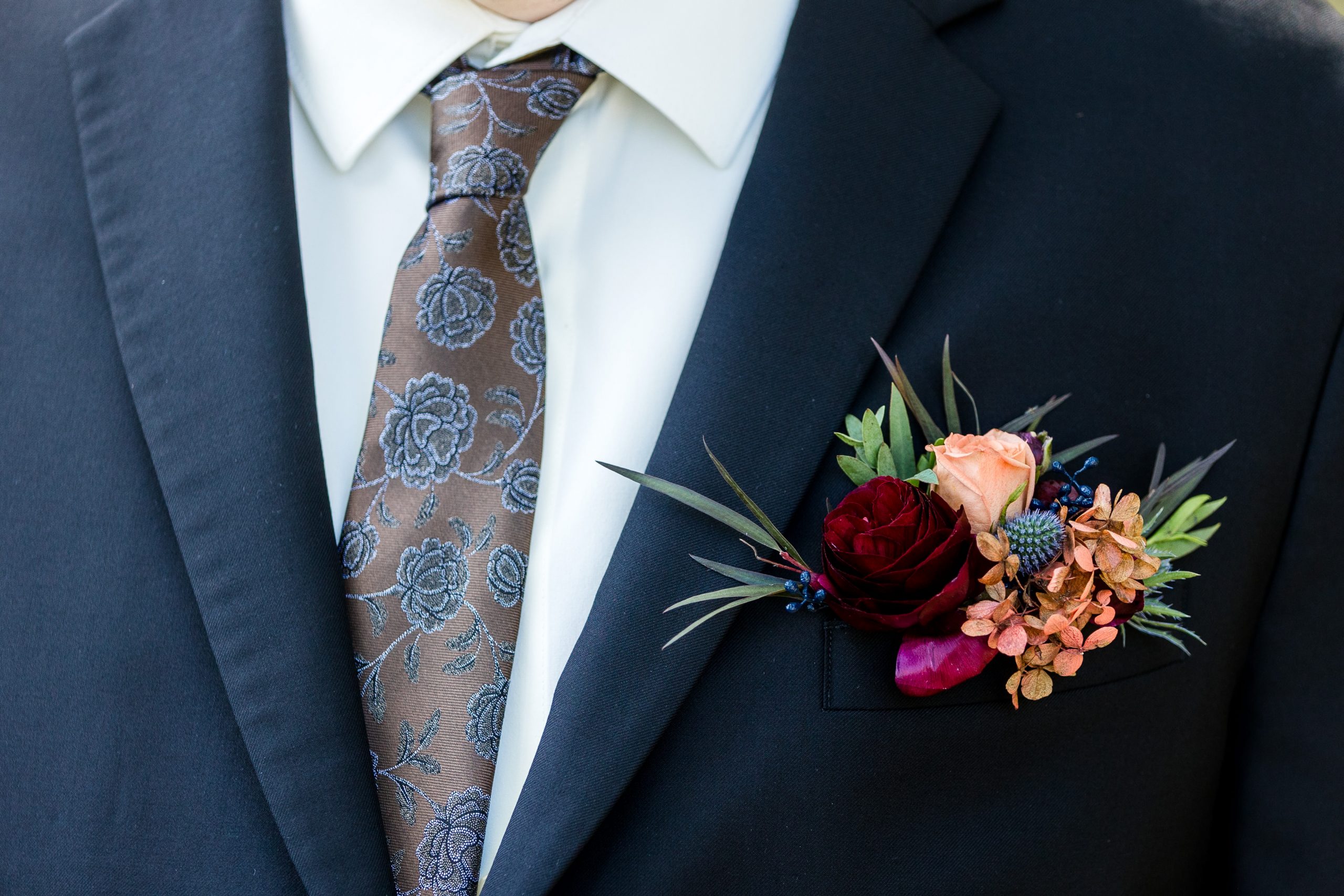 Close-up of a groom’s suit with a floral boutonniere featuring peach, burgundy, and rust-colored blooms.