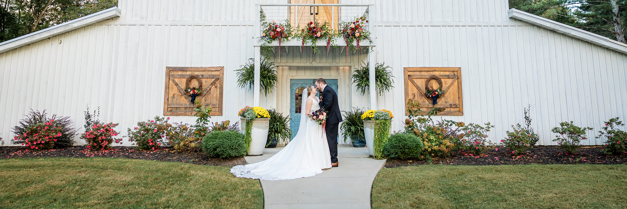 Bride and groom sharing a kiss outside a white barn wedding venue in Huntsville, decorated with colorful floral arrangements.